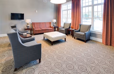 A well-lit living room area with a patterned carpet, a red sofa, two gray armchairs, a white ottoman in the center, side tables with lamps, and large windows with red curtains allowing natural light to enter.