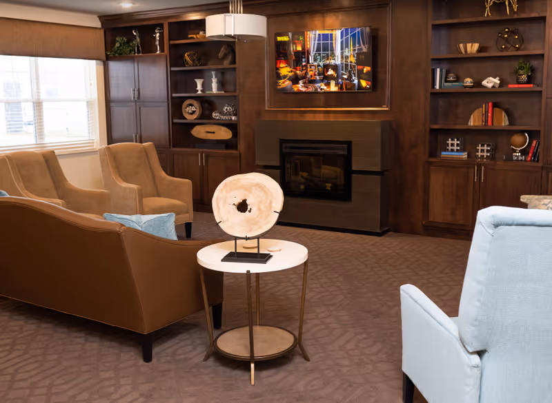 A cozy living room with a brown leather sofa, two beige armchairs, and a light blue armchair. A small round table with a decorative wooden piece is in the center. The room features built-in dark wood shelves and cabinets, a fireplace, and a large window with blinds.