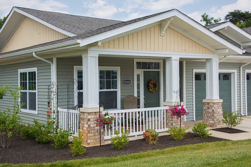 Exterior view of a single-story residential unit at Traditions at Beaumont featuring a covered front porch with white railings and brick pillars, a green front door decorated with a floral wreath, two windows with white trim, hanging flower baskets, and a well-maintained garden with shrubs and flowers.