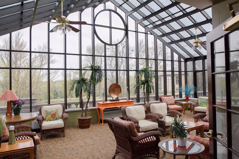 A bright sunroom with large floor-to-ceiling windows and a glass ceiling, furnished with wicker chairs and sofas with cushions, small tables with plants and lamps, and ceiling fans. Outside, trees and greenery are visible through the windows.