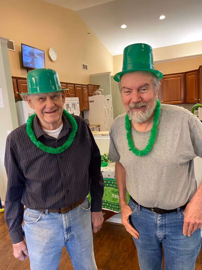 Two elderly men standing indoors in a kitchen area, both wearing green plastic hats and green leis, smiling and posing for the photo. The kitchen has wooden cabinets, a refrigerator, and a table with a green St. Patrick's Day themed tablecloth in the background.