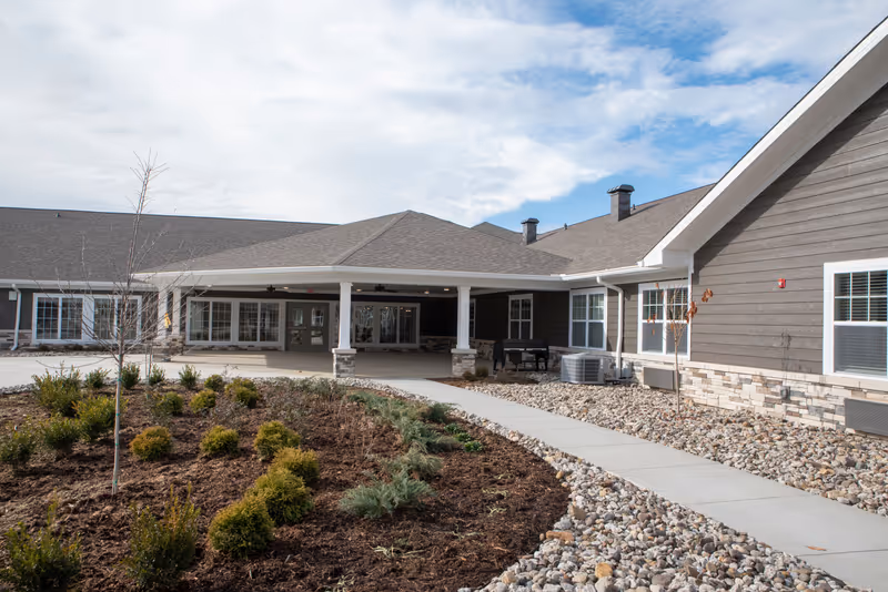 Exterior view of Cooper Trail Senior Living building with a covered entrance, landscaped garden with small bushes and young trees, a concrete walkway, and a partly cloudy sky.