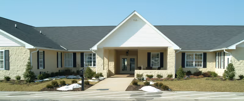 Front exterior view of a single-story assisted living facility building with beige walls, black shutters, a covered entrance with double glass doors, and a small landscaped area with bushes and patches of snow.