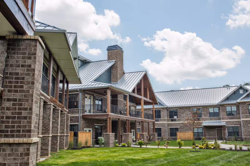 Exterior view of a senior living facility with brick walls, metal roofs, balconies, and a well-maintained lawn under a partly cloudy sky.