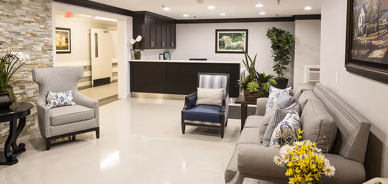 A well-lit reception area with a dark wood front desk, a gray sofa with patterned pillows, a blue armchair, and a gray wingback chair. There are several potted plants and flowers, a stone accent wall, and framed artwork on the walls.
