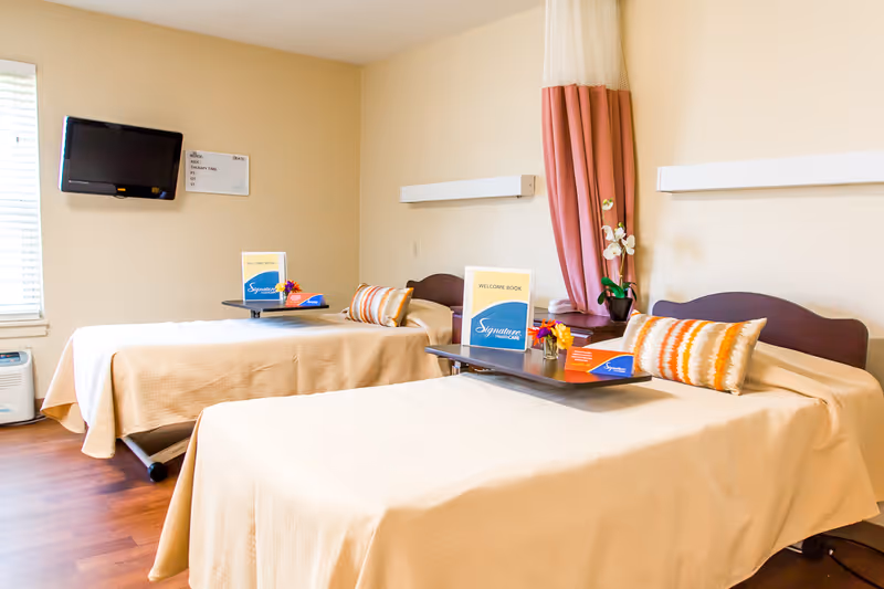 A bright and clean bedroom with two single beds covered in beige bedspreads, each with a decorative pillow and a small table holding a welcome book and flowers. The room has a wall-mounted TV, a window with blinds, and a pink curtain partially drawn to the side.