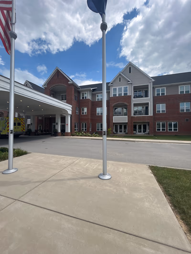 Front exterior of a three-story brick senior living building with a covered entrance, flagpoles, and an ambulance parked near the porte-cochère.