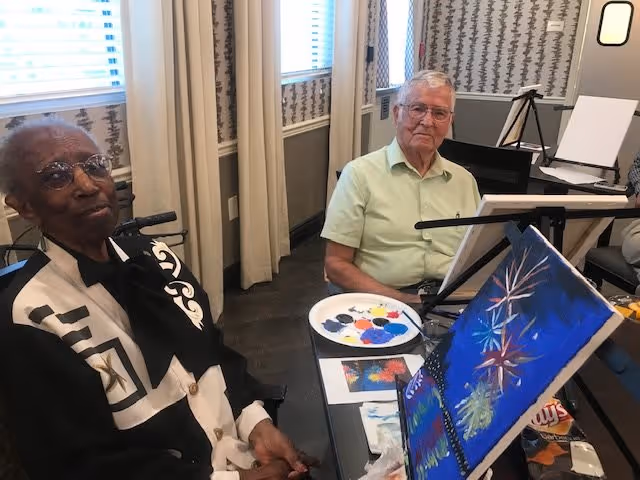 Two elderly individuals seated at a table in a room with windows and curtains. One person is wearing glasses and a patterned jacket, while the other is wearing a light green shirt. On the table in front of them are painting supplies, including a palette with paint and a canvas with a colorful painting featuring fireworks.