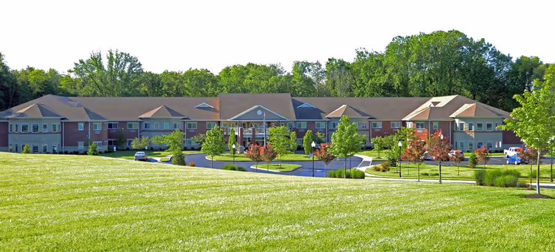Wide exterior view of Magnolia Springs East Louisville, a large two-story senior living facility surrounded by green grass, trees, and a circular driveway with parked cars.