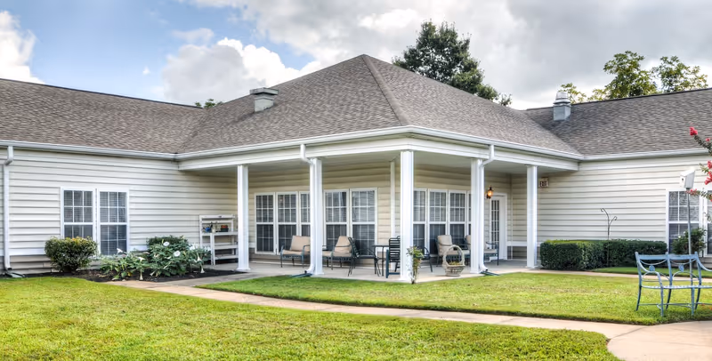 Exterior view of a single-story senior living facility building with beige siding and a gray shingled roof. The building features a covered patio area with several chairs and small tables. There are well-maintained green lawns and shrubs surrounding the building under a partly cloudy sky.