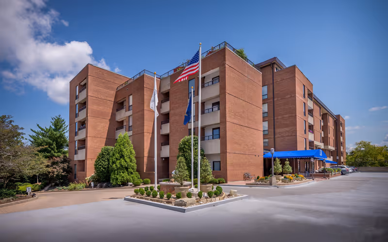 Exterior view of a multi-story brick building with balconies, three flagpoles with flags in front, landscaped greenery, and a blue awning over the entrance under a partly cloudy sky.