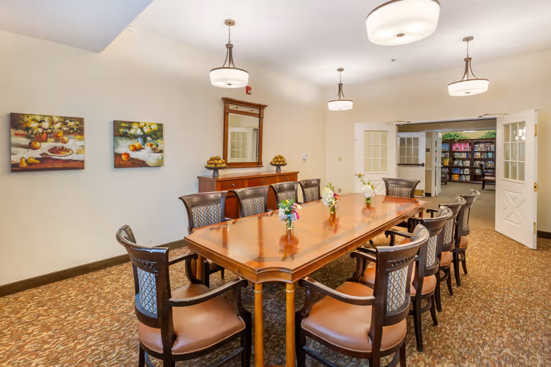 A well-lit dining room with a long wooden table surrounded by ten chairs. The table has small flower arrangements placed along its center. The room features beige walls, two paintings of fruit on the left wall, a wooden sideboard with a mirror above it, and double doors leading to a room with bookshelves.