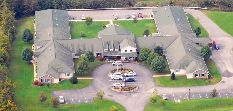 Aerial view of a large single-story building with a U-shaped layout surrounded by green lawns and trees. The building has a gray roof and beige walls, with a parking lot in front containing several cars. The area is bordered by a road and additional greenery.