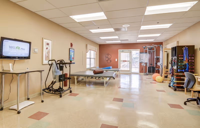 A spacious physical therapy or exercise room with beige walls and a tiled floor. The room contains exercise equipment including a multi-gym machine, therapy tables with bolster pillows, exercise balls, and racks with various weights and resistance bands. A flat-screen TV mounted on the wall displays the Wii Fit Plus game. There are windows allowing natural light and a door leading outside.