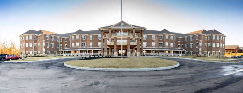 Front exterior view of a large, three-story brick senior living facility with multiple windows, a central entrance with columns, and a circular driveway with a grassy island in the middle.
