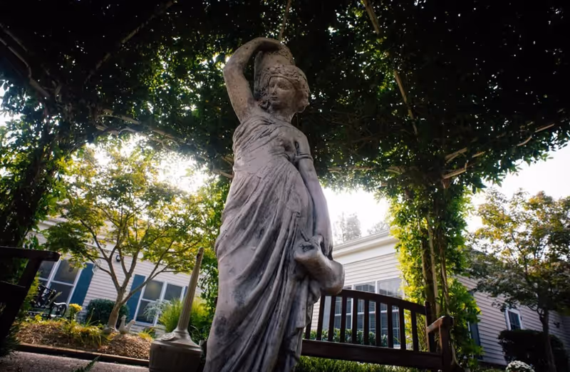 Stone statue in a shaded garden courtyard at Gaither Suites with bench and trees.