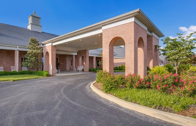 Exterior view of Inspirations of Mount Washington facility showing a brick building with a covered entrance, a driveway, green shrubs, flowering plants, and a clear blue sky.
