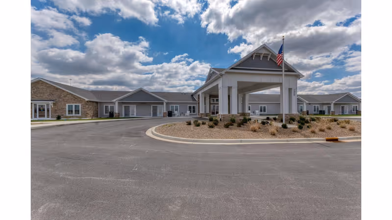 Front entrance of a single-story senior living facility with a porte-cochere, American flag, and circular driveway under a partly cloudy sky.