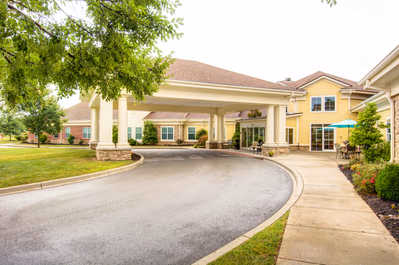Entrance of a senior living facility with a covered driveway supported by columns, surrounded by well-maintained landscaping and outdoor seating with an umbrella.