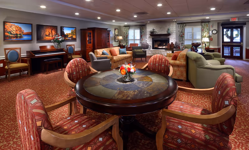 A cozy senior living facility common area with a round table and four patterned chairs in the foreground. The room features multiple sofas and armchairs arranged around a stone fireplace with a fire burning. There is a piano against the wall with framed autumn landscape paintings above it. The room is warmly lit with recessed ceiling lights and lamps, and has patterned carpet and floral curtains on the windows. An exit door is visible in the background.