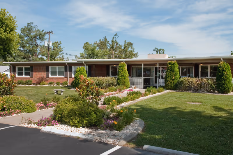 Single-story brick building with a covered entrance, surrounded by well-maintained green lawns, shrubs, and flowering plants under a partly cloudy sky.