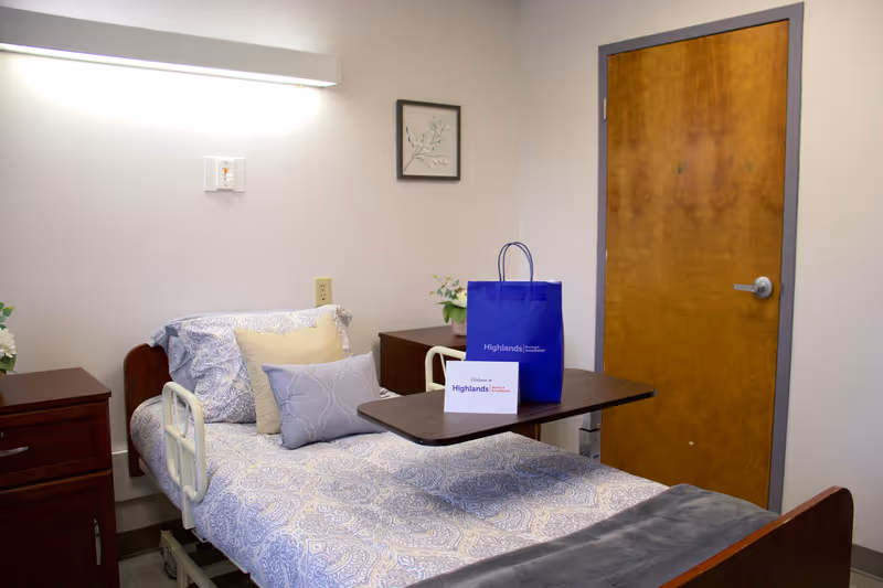 Hospital-style patient room with a bed, blue bedding, and a closed wooden door.
