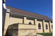 Exterior view of a beige brick building with a brown roof under a clear blue sky. The building features tall, narrow, arched windows and a stone base wall in the foreground.