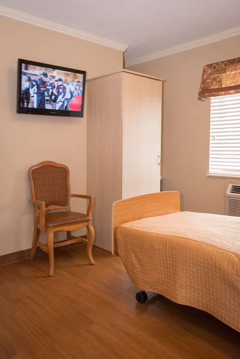 A simple bedroom in a senior living facility featuring a single bed with a beige quilt, a wooden chair with patterned upholstery, a light wood wardrobe, a wall-mounted flat screen TV showing a scene with people, and a window with blinds and a decorative valance.