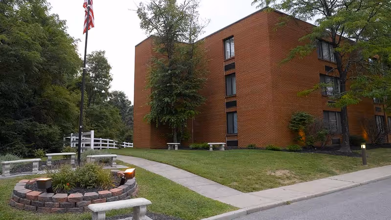 Exterior view of a multi-story brick building with several windows, surrounded by green grass, trees, and a paved walkway. There is a circular brick planter with plants and lights, and an American flag on a flagpole. Stone benches are placed around the planter and along the walkway.