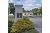 Stone entrance pillar with a Fern Terrace sign beside shrubs and a driveway leading into the facility grounds under a blue sky.