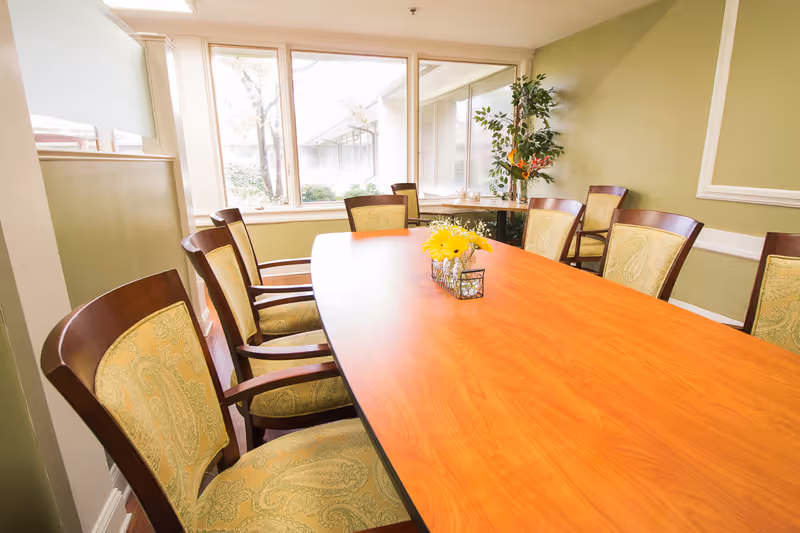 Long wooden table with a small floral centerpiece surrounded by upholstered chairs in a sunlit room with green walls and large windows.