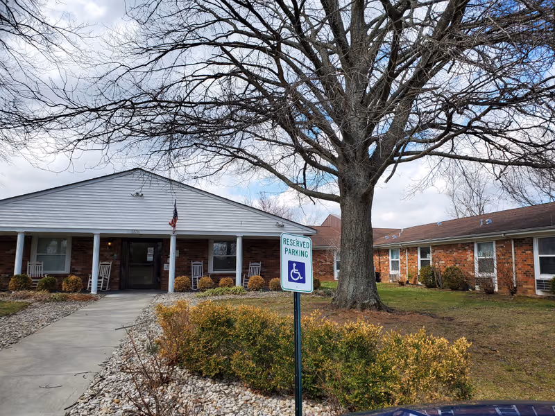 Single-story brick senior living building with a covered entrance, tree, and handicap parking.