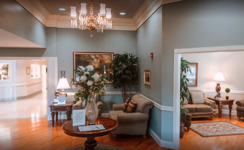 A warmly decorated nursing facility sitting area with a round table topped by a vase of flowers, upholstered chairs, lamps, and a chandelier.