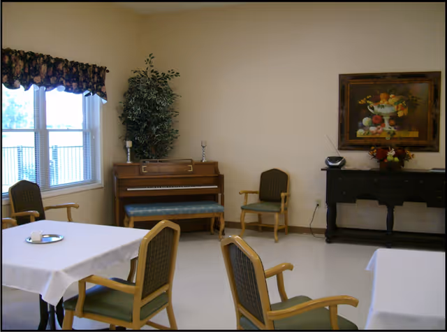 A common area with several tables covered in white tablecloths and surrounded by wooden chairs with green cushions. A piano with a blue cushioned bench is against the wall, next to a tall artificial plant. On the right side, there is a black console table with a floral arrangement and a small radio, above which hangs a framed painting of a fruit bowl. A window with floral valance lets in natural light.