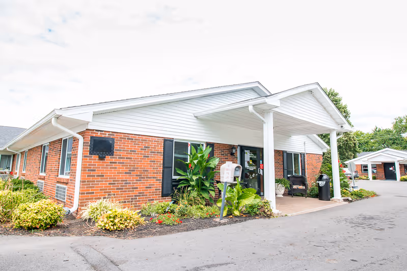 Exterior view of a single-story brick building with white siding and a covered entrance supported by white columns. There are plants and shrubs along the building's foundation, a mailbox near the entrance, and a paved driveway in front. The sky is partly cloudy.