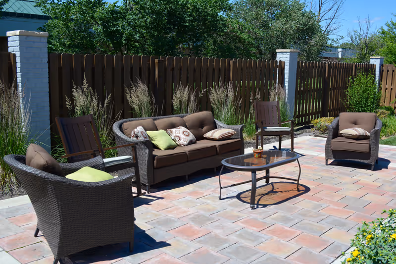 Outdoor patio area with brown wicker furniture including a sofa, two armchairs, and two wooden chairs arranged around a glass-top coffee table. The patio is paved with multicolored bricks and is bordered by a wooden fence with white brick pillars and green plants.