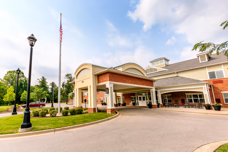 Covered entrance of a brick senior living building with a flagpole, driveway, and landscaping.