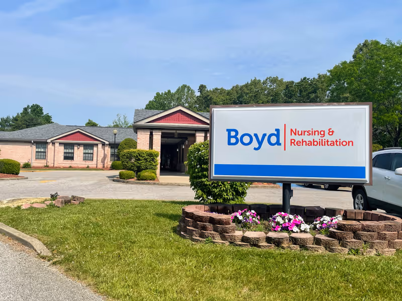 Exterior view of Boyd Nursing and Rehabilitation facility with a large sign in the foreground displaying the facility's name. The building is a single-story brick structure with a covered entrance, surrounded by greenery and a parking area with a white vehicle visible.