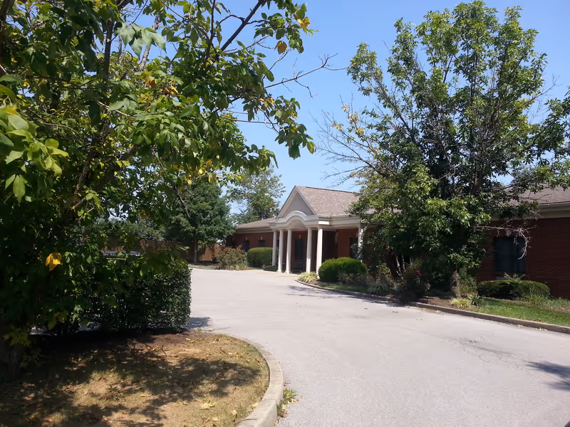 Brick senior living facility entrance with columns, a driveway, and surrounding trees.