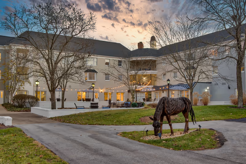 Exterior view of a senior living facility at dusk with a three-story building, leafless trees, string lights, outdoor seating area, and a metal sculpture of a horse grazing on a small patch of grass.