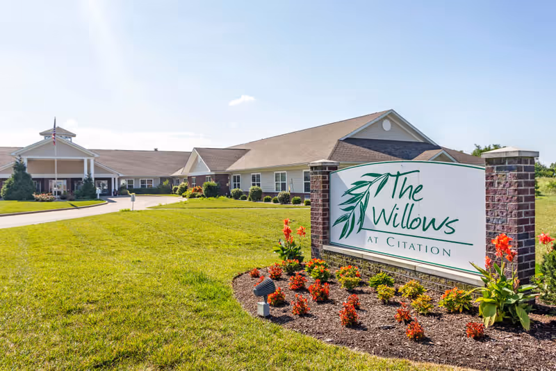 Exterior view of The Willows at Citation senior living facility with a large sign in the foreground surrounded by flowers and a well-maintained lawn, and the building visible in the background under a clear blue sky.