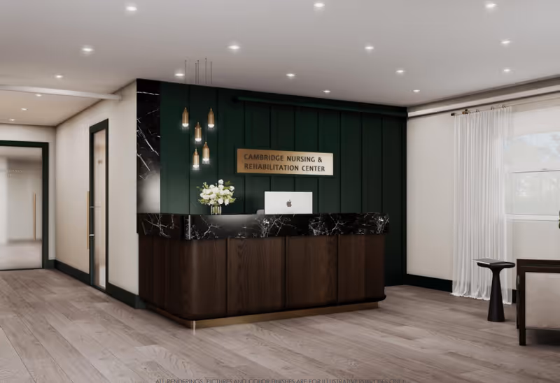 Reception area of Cambridge Nursing & Rehabilitation Center featuring a dark wood and black marble reception desk with a computer and a vase of white flowers. Behind the desk is a dark green paneled wall with a sign displaying the facility's name. The area has light wood flooring, pendant lights hanging above the desk, and a window with sheer white curtains on the right side.