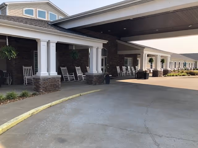 Covered entrance area of a senior living facility with white columns, stone bases, hanging plants, and several white rocking chairs lined up along the porch.