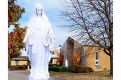 A large white statue of a religious figure, likely the Virgin Mary, stands outdoors in front of a brick building with leafless trees and some greenery around.
