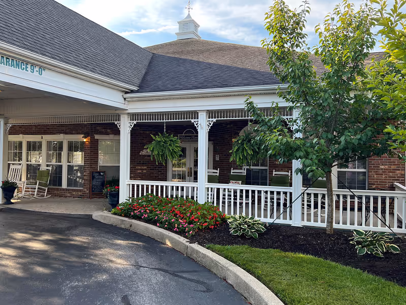 Exterior view of Morning Pointe of Richmond showing a covered entrance with a white railing and brick walls. There are hanging plants, rocking chairs, a small garden with flowers and shrubs, and a sign that reads 'Welcome Home' above the door.
