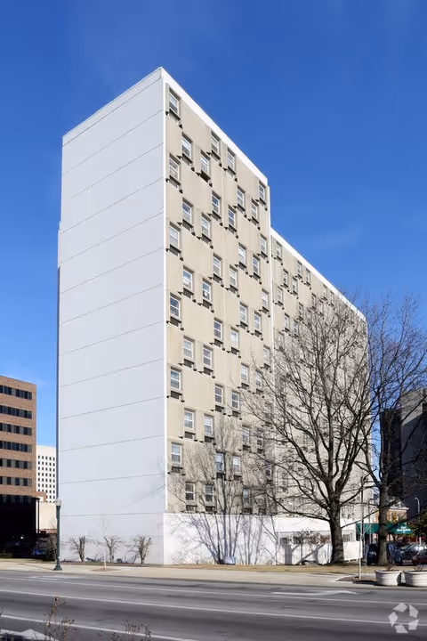 Tall multi-story beige and white building with many windows, situated next to a street with leafless trees and a clear blue sky in the background.