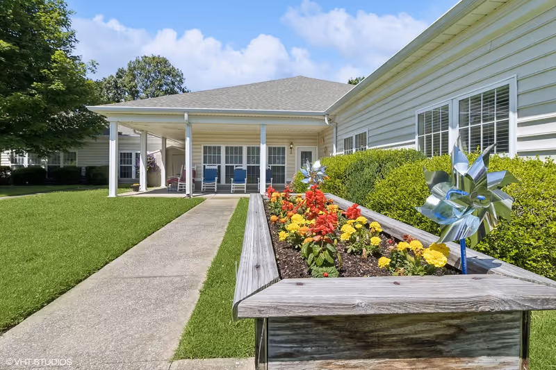 Front exterior of a single-story building with a covered porch, walkway, and a raised flower planter with pinwheels.