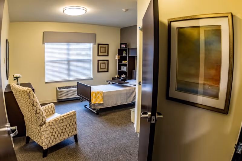 A furnished single bedroom with a bed, armchair, window and framed artwork seen through an open door.