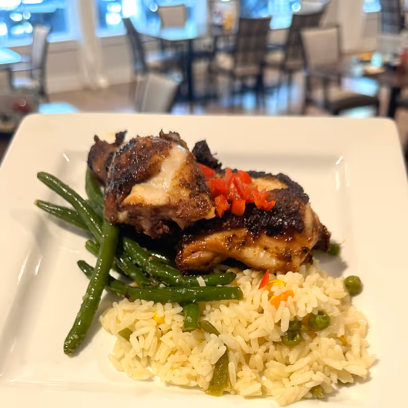 Grilled chicken with green beans and seasoned rice served on a square white plate in a dining room setting.