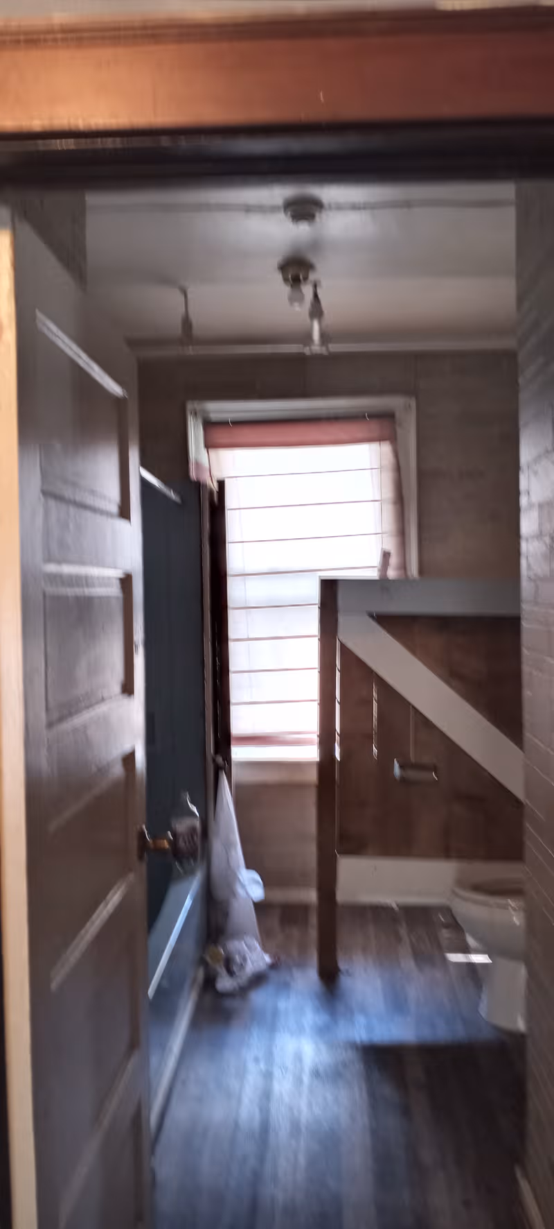 Narrow bathroom with toilet, window, and wooden railing in a dim hallway.